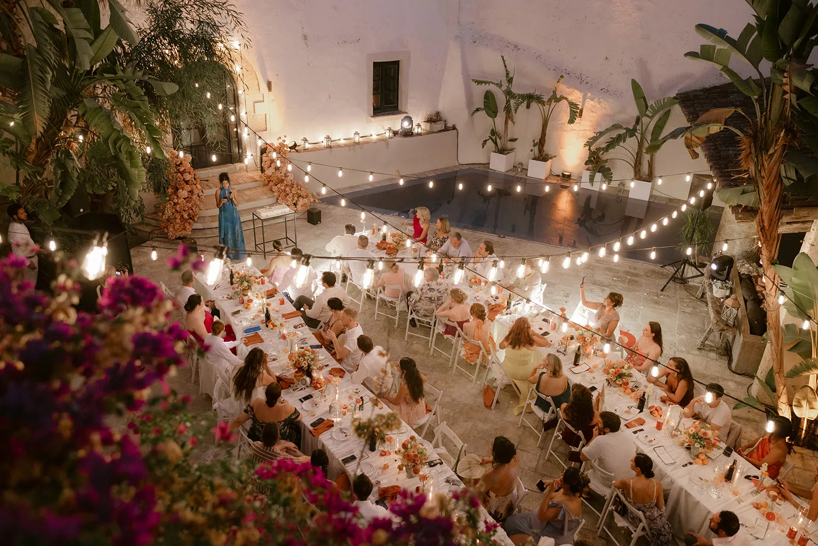 Wedding guests seated at long tables in the courtyard under festoon lights
